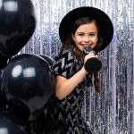 portrait of a young brunette singer in a black dress singing with a microphone in her hands on a shiny background in helium balloons