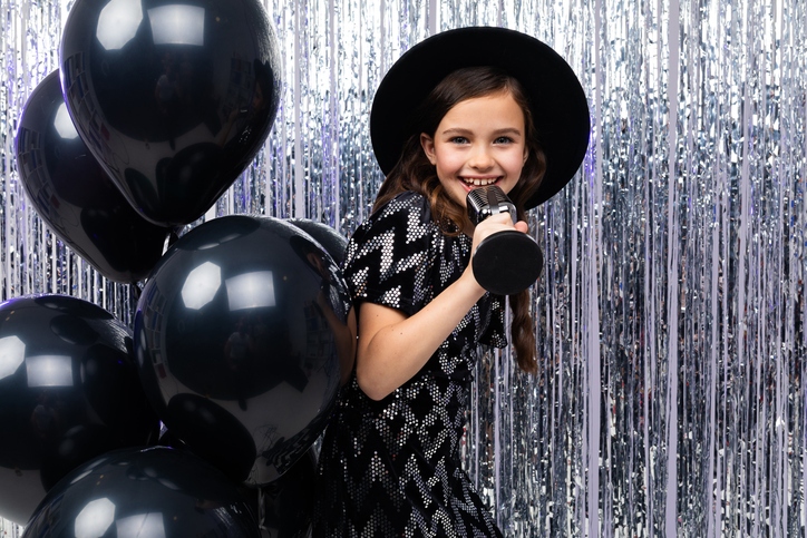 portrait of a young brunette singer in a black dress singing with a microphone in her hands on a shiny background in helium balloons
